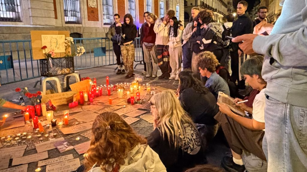 People gather around a vigil where red candles and written tributes sit on pavement near a fence.