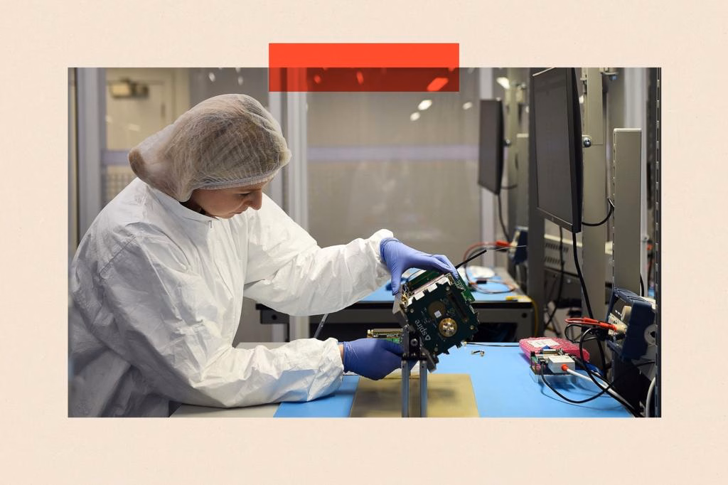 Satellite manufacturing technician Debbie Wardhaugh works at the offices of US satellite firm Spire Global in Glasgow. She is wearing a hairnet, white overalls and blue rubber gloves and is making adjustments to a CubeSat device. In front of her is a row of screens.