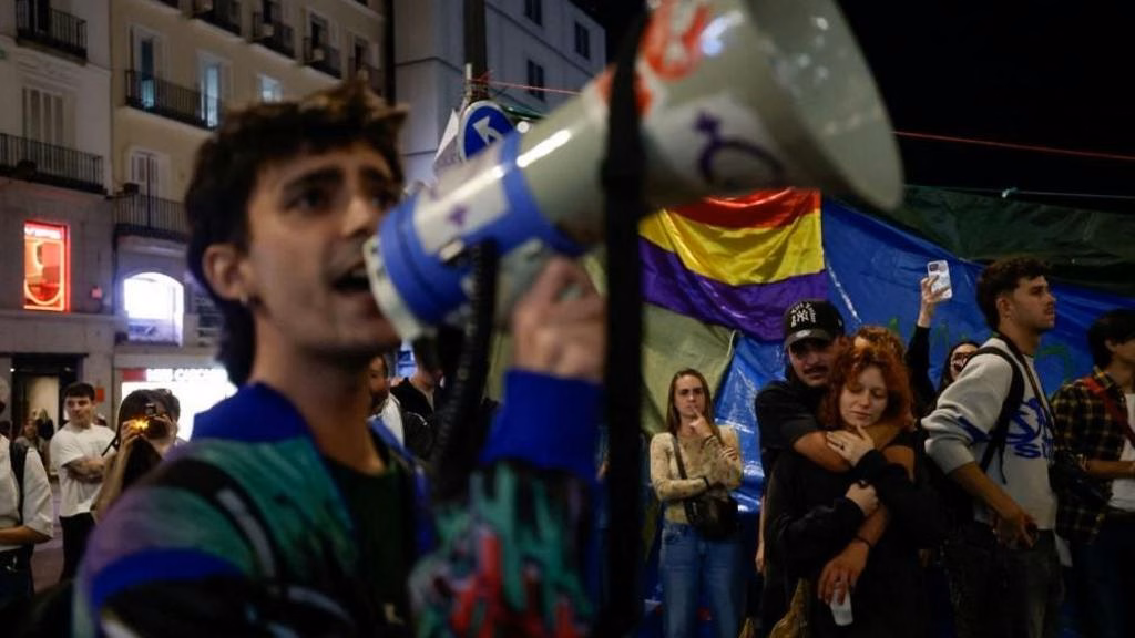 A demonstrator speaks into a megaphone in Puerta del Sol, Madrid. Two protesters are stood behind him hugging.